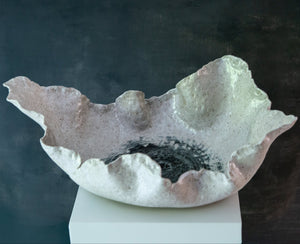 An organic-shaped ceramic bowl with a textured, speckled finish in off-white, beige, featuring a dark charcoal center, displayed on a white pedestal against a dark background.