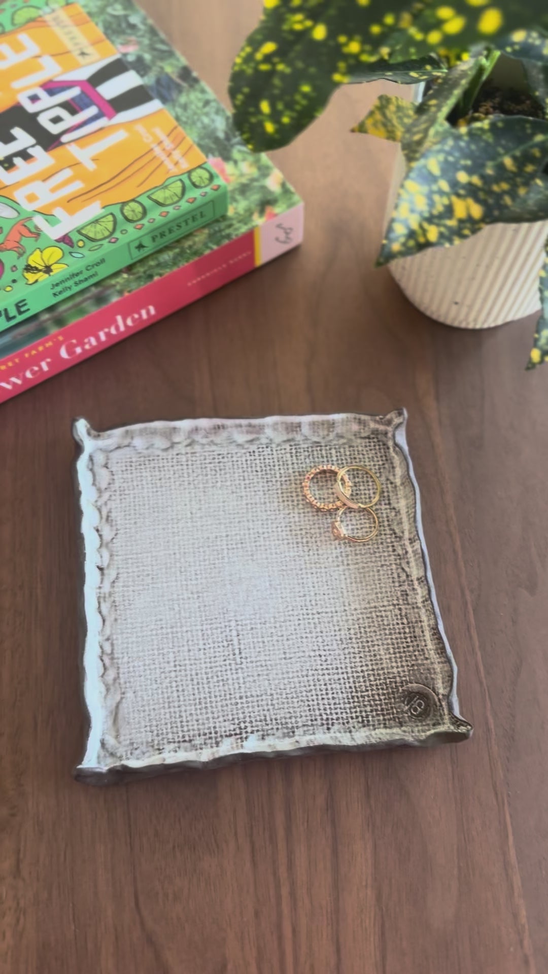 Gold hoop earrings on a fabric pad with a hand touching it, on a wooden surface with books and a plant in the background.